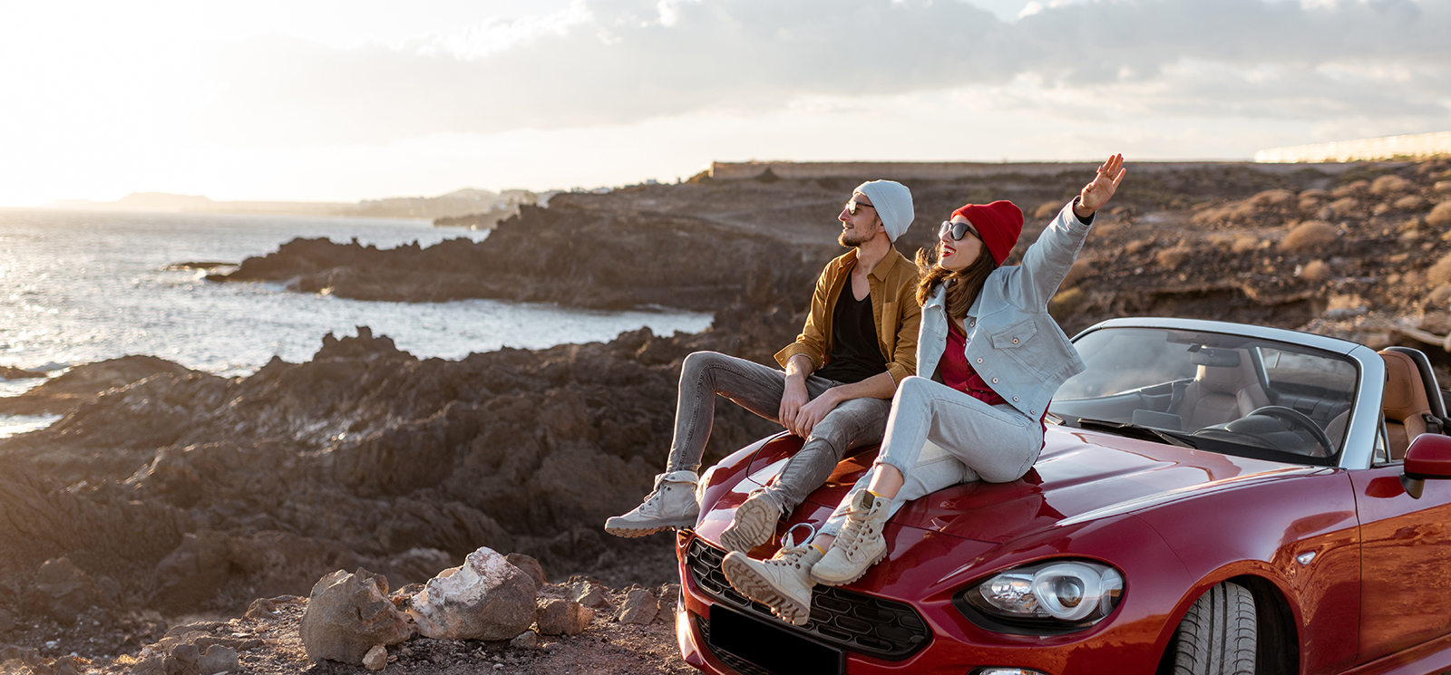 Couple sitting on hood of red convertible on mountain near the ocean